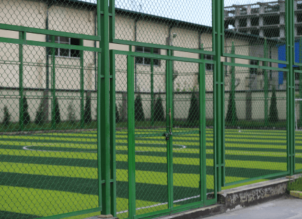 An empty green outdoor paddle tennis court enclosed by a tall green metal fence, with a cream-colored building visible in the background, owned by Tajhind Edutech Pvt Ltd.